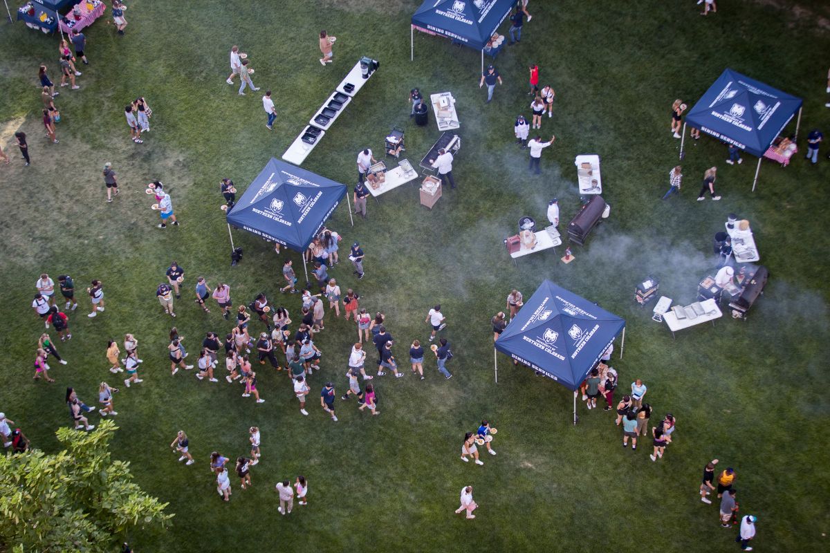 A large crowd of students gathers on a grassy field with multiple blue UNC Dining Services tents and barbecue grills set up, smoke rising as food is being served.
