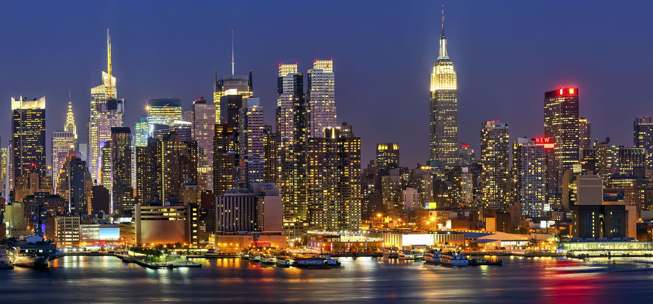 A vibrant nighttime cityscape of New York City featuring illuminated skyscrapers, including the Empire State Building, reflecting off the adjacent water.
