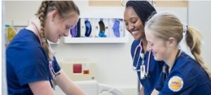Three nursing students are at a hospital bed in a learning lab. They are wearing blue scrubs and have stethoscopes around their necks.