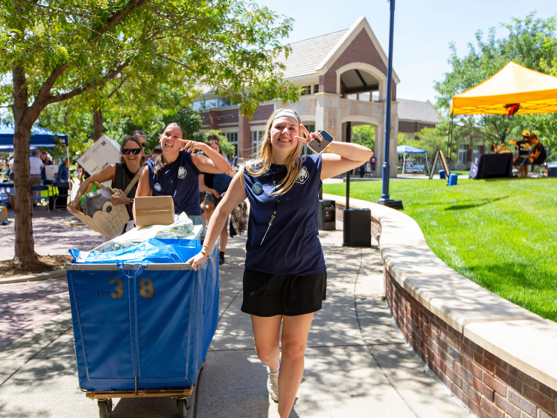 UNC student volunteers smile and flash peace signs while helping move belongings in large blue carts during Move-In Day.