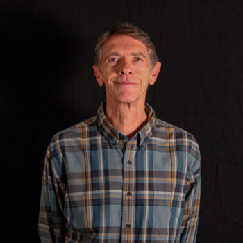 A man smiling, dressed in a flannel, against a black background