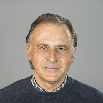 A man smiling, dressed in a blue sweater. Against a light grey background.