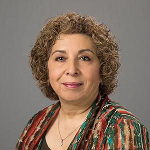 A woman smiling, dressed in a colorful sweater, against a grey background.