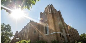 This photo shows Gunter Hall, a tan brick building on campus, with blue sky, green trees, and sun beams in the background.