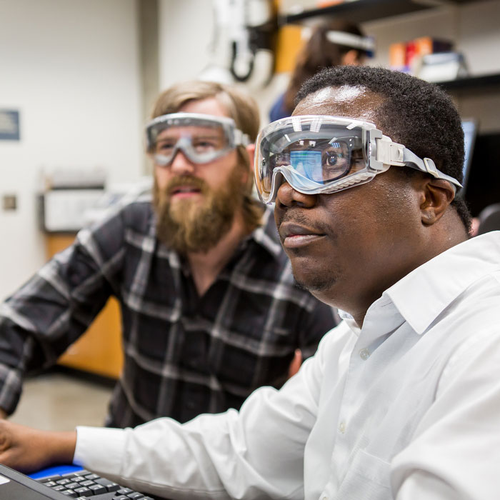Two chemistry graduate students wearing safety goggles work together on data analysis at a computer in a campus lab.