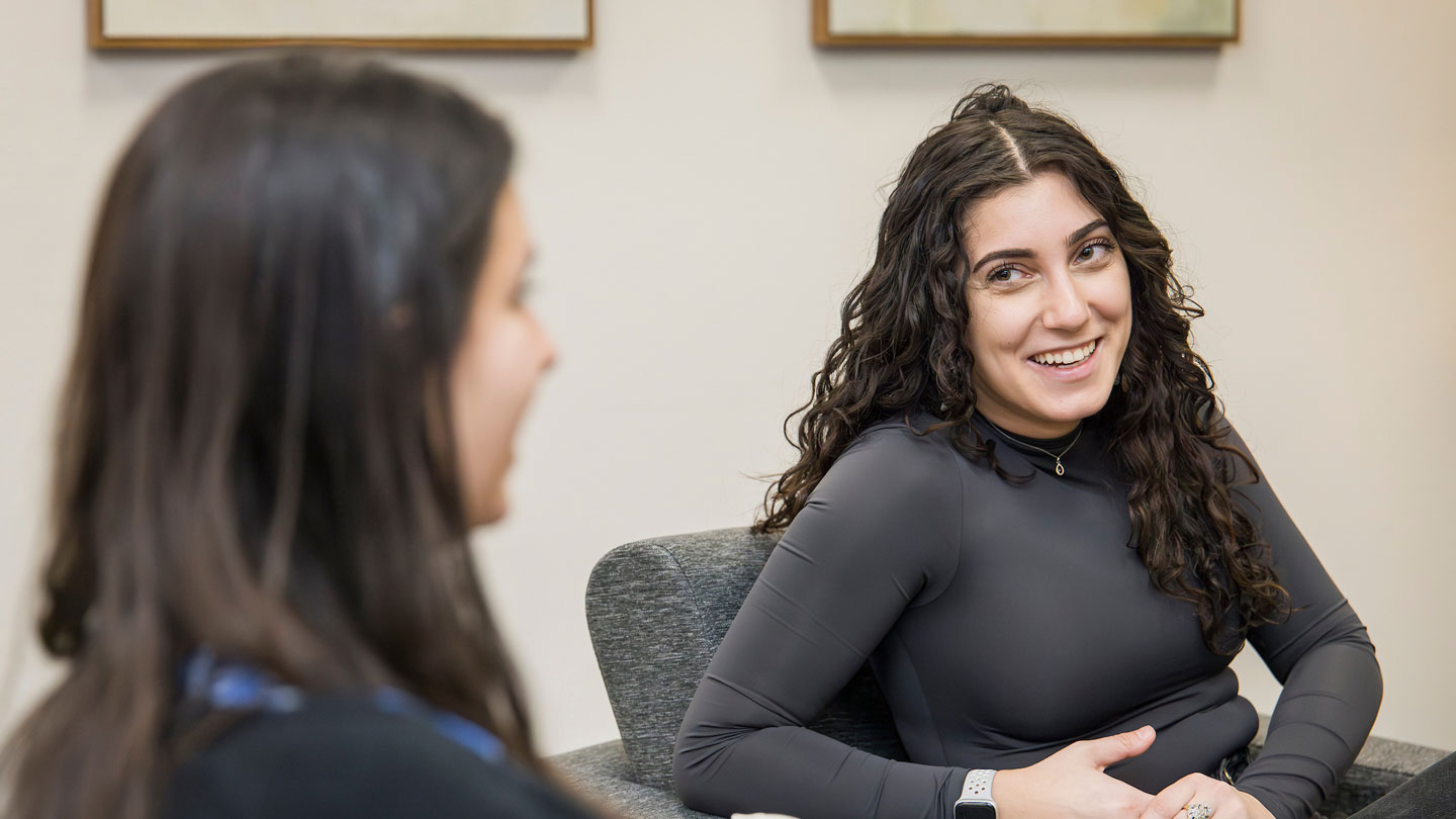 Student smiling while listening to another student talk in classroom setting.