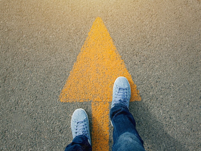 Feet of man in sneakers walking across yellow arrow on street.