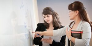 A professor and a student are standing next to a white board. The professor is pointing out somethin on the board while the student writes in a notebook.