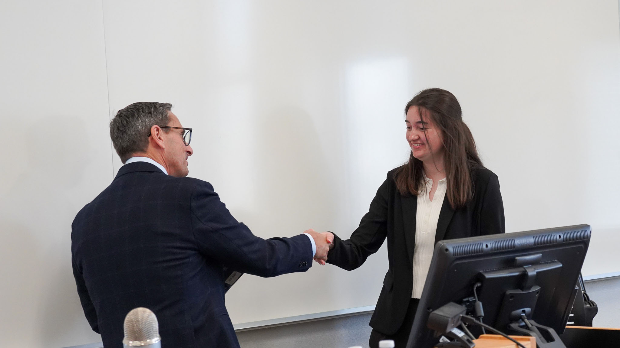 A young woman in a black blazer and white blouse is shaking hands with a man in a dark suit and glasses in a classroom or conference room setting.