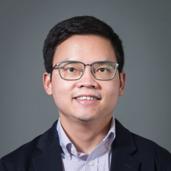 A man with glasses smiling, wearing a dark blue suit jacket, against a dark grey background.