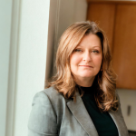 A woman smiling, with light hair, dressed in a grey blazer. 