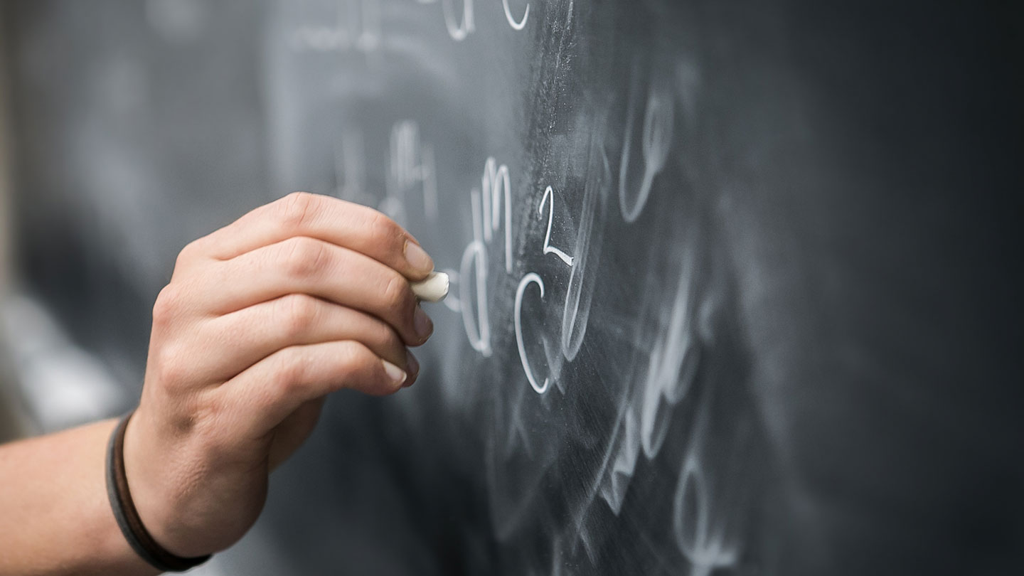 Hand, with chalk, writing an equation on a chalkboard.