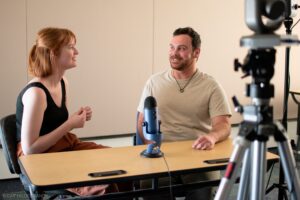 Two students sitting behind a table in the Michener Library Recording Room. There is a Yeti microphone on the table and a camera on a tripod in the foreground.