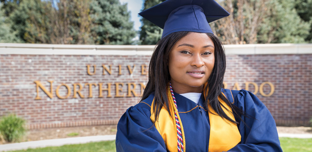 Sandra Mbayo wearing her graduation cap and gown and posing for the camera in front of a UNC brick sign