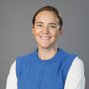 A person smiling, wearing a blue sweater over a white shirt, set against a neutral background.