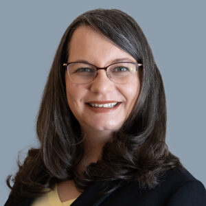 A woman with dark hair, glasses and dressed in a black suit. Smiling for a photo 