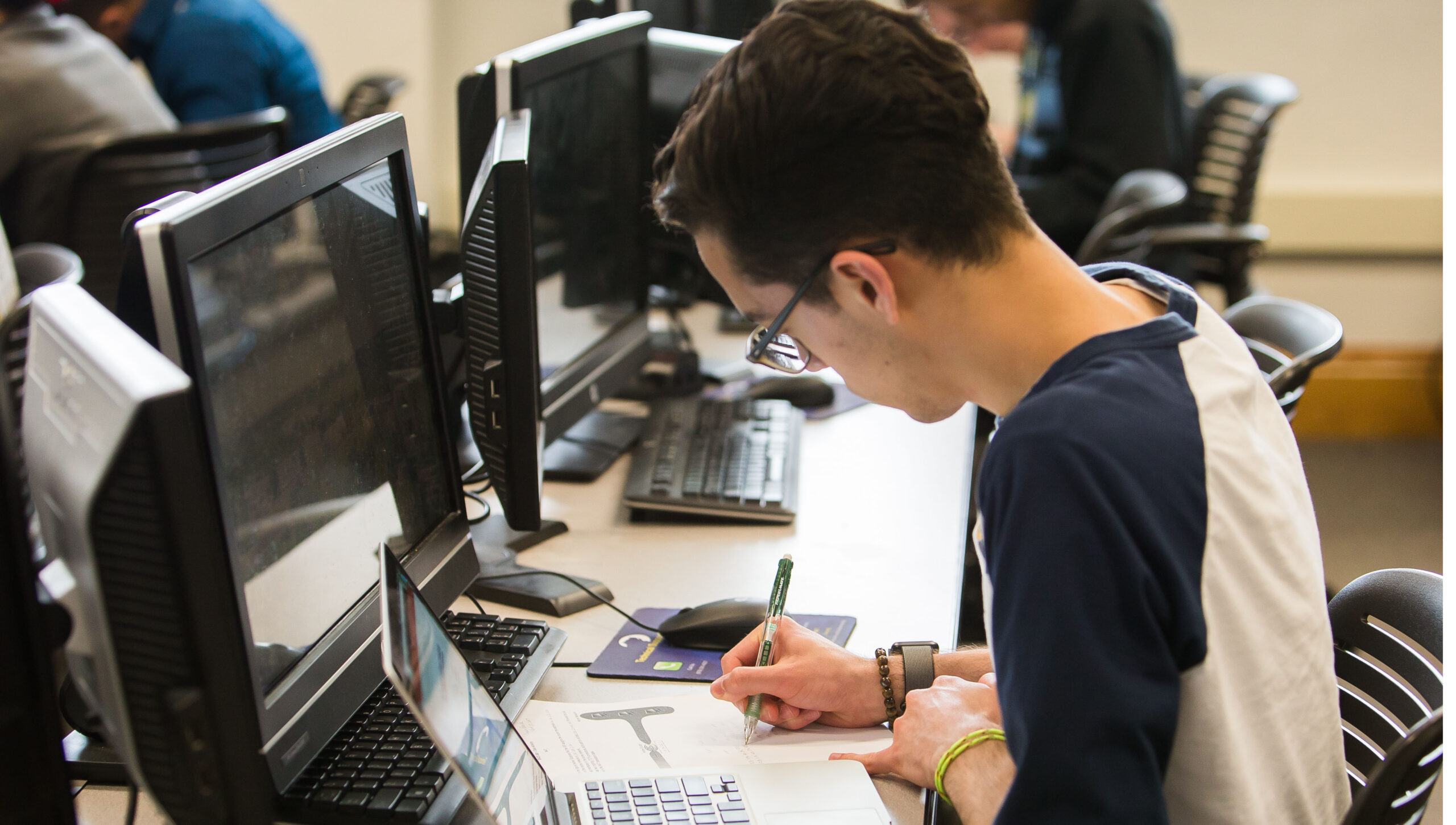A student focuses on writing notes while seated at a computer workstation in a classroom setting, surrounded by monitors and other students.