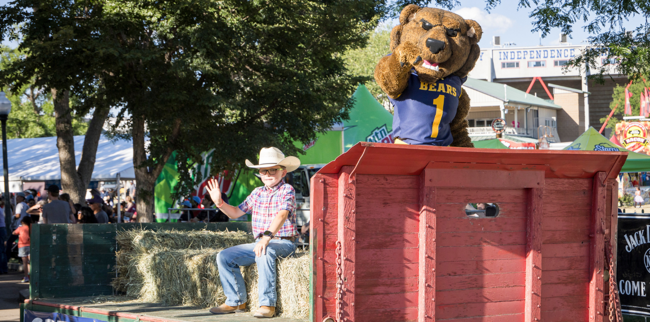 A kid wearing a cowboy hat sitting on hay during a parade with Klawz standing in the front pointing