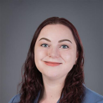 A woman with red hair, smiling, against a dark grey background