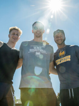 A group of three male students, looking down, smiling, the sun is shining near their faces.