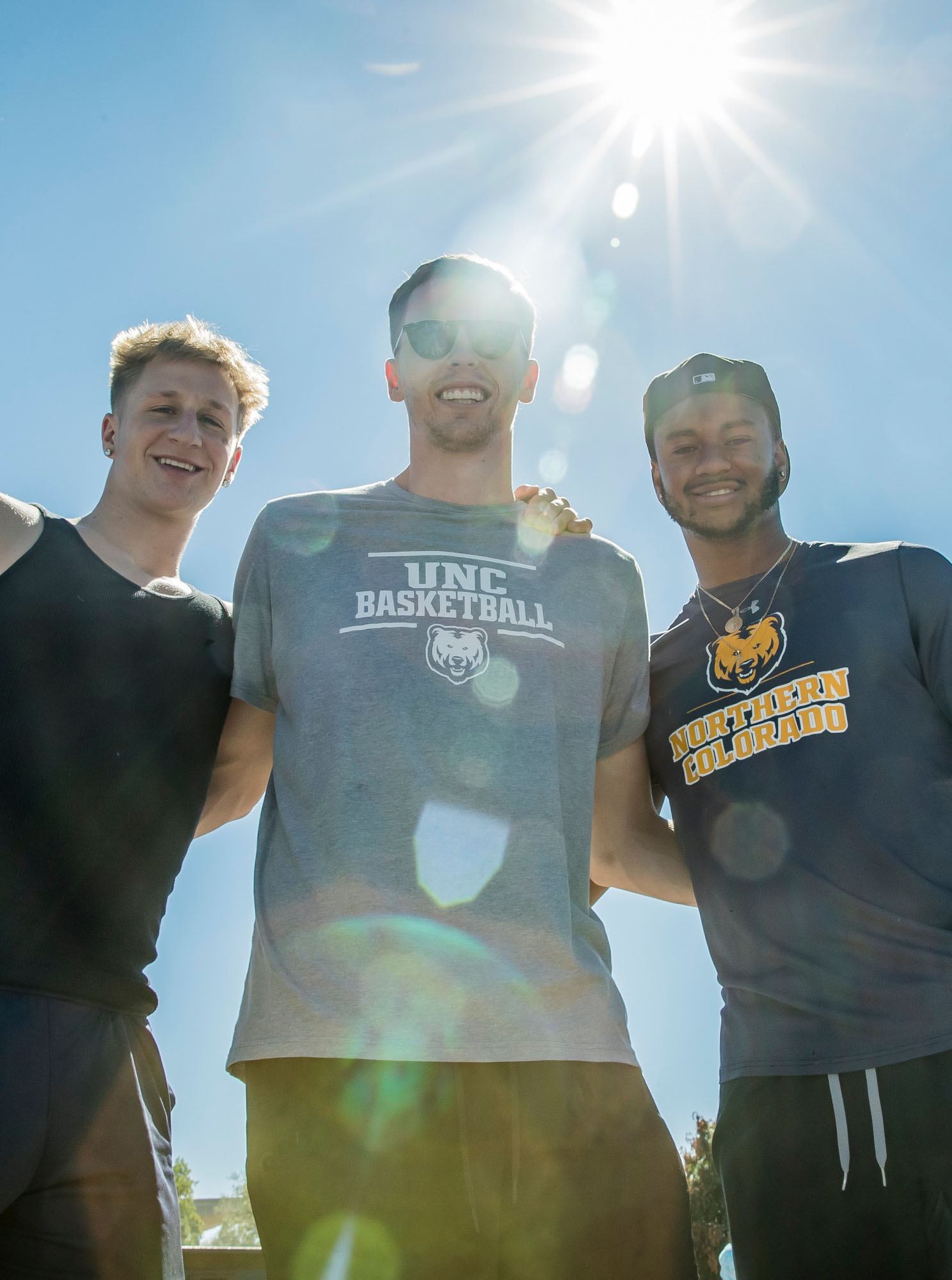 A group of three male students, looking down, smiling, the sun is shining near their faces.