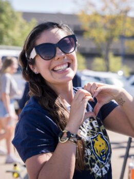 A woman with long brown hair, wearing large dark sunglasses, smiling brightly and making a heart shape with her hands. She is dressed in a navy blue t-shirt with a lion logo, and has a tattoo of a flower on her wrist. The background shows a sunny outdoor setting with blurred people and trees.