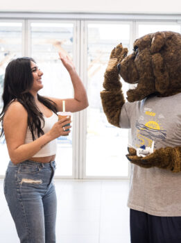 A young woman with long dark hair, wearing a white crop top and jeans, sharing a high-five with a Klawz the bear