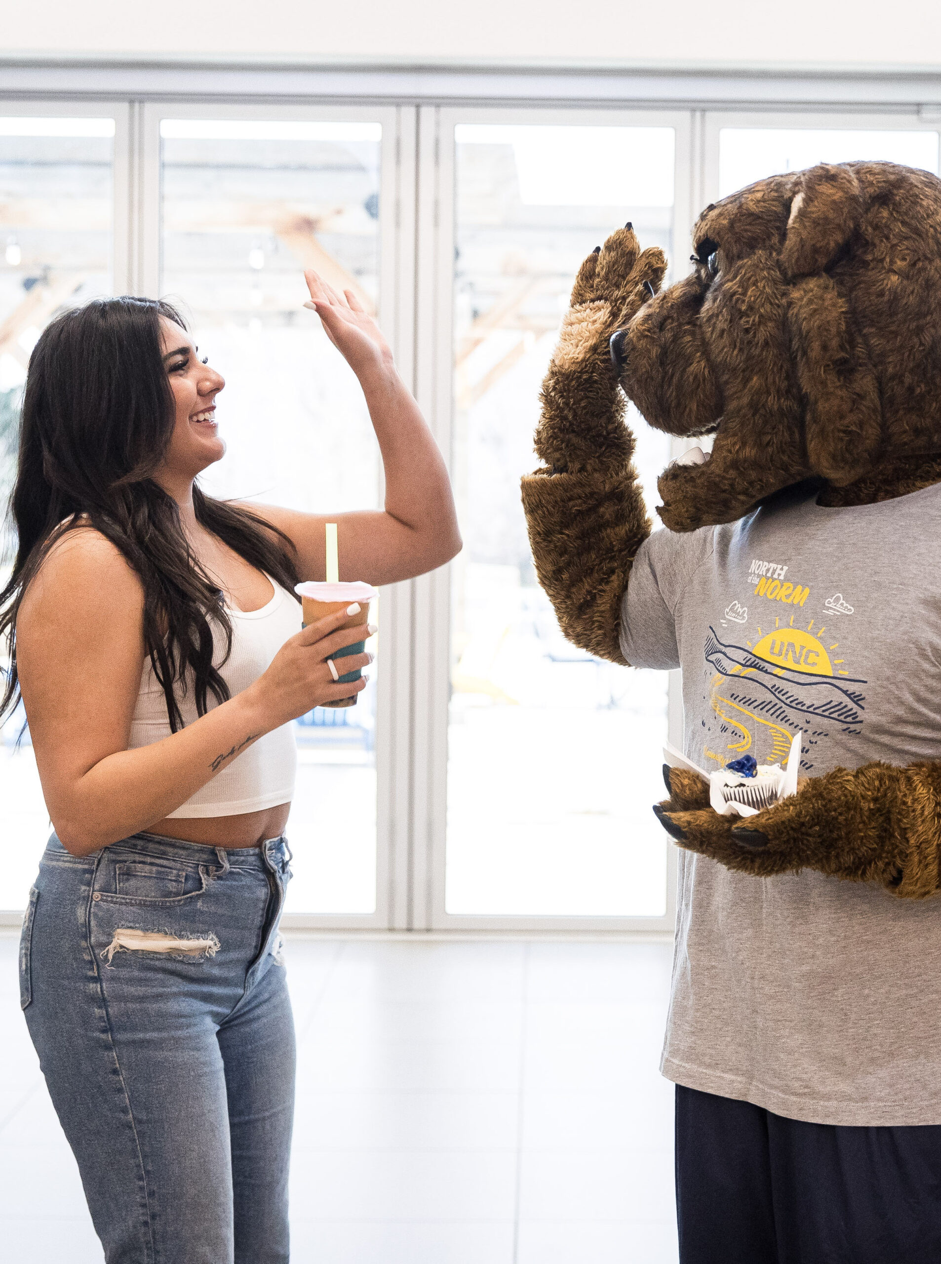 A young woman with long dark hair, wearing a white crop top and jeans, sharing a high-five with a Klawz the bear