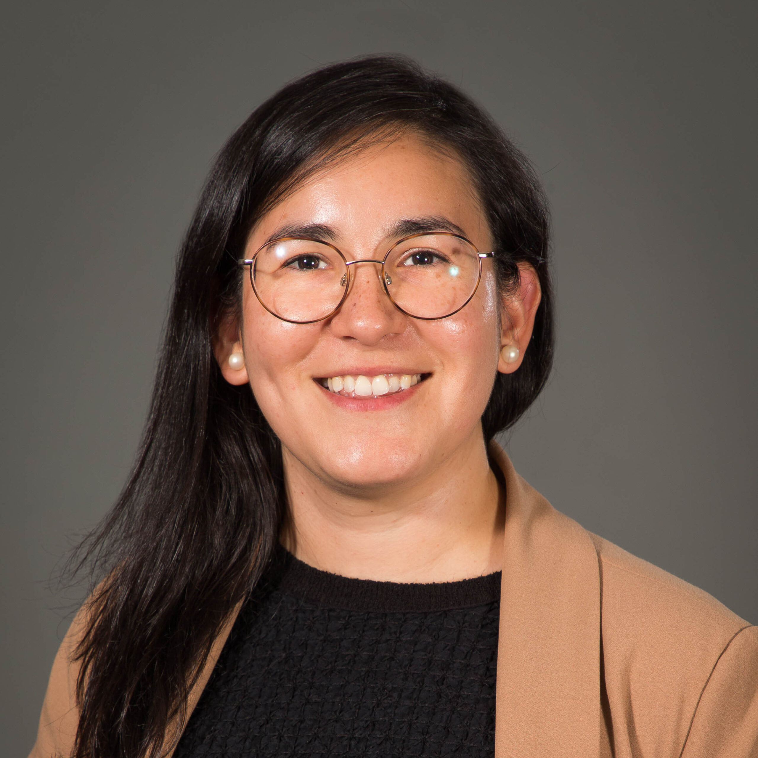 A woman with dark black hair, dressed in a tan jacket, against a dark grey background.