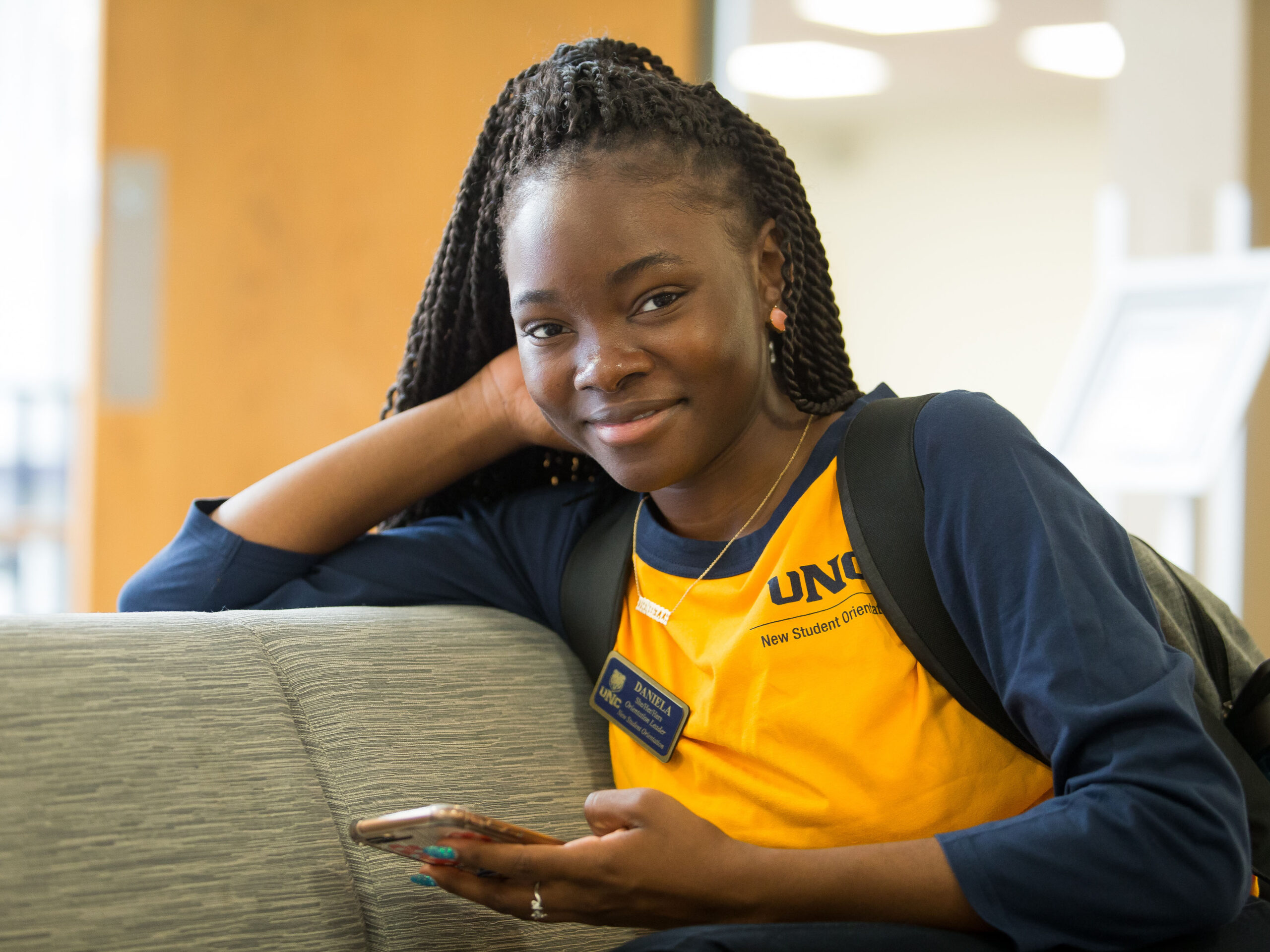 A young woman with braided hair, smiling and relaxing on a couch while holding a smartphone. She is wearing a yellow and navy blue shirt with a name tag and a backpack.