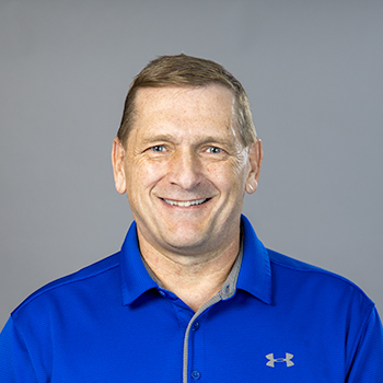 A man smiling, dressed in a blue polo shirt, against a grey background