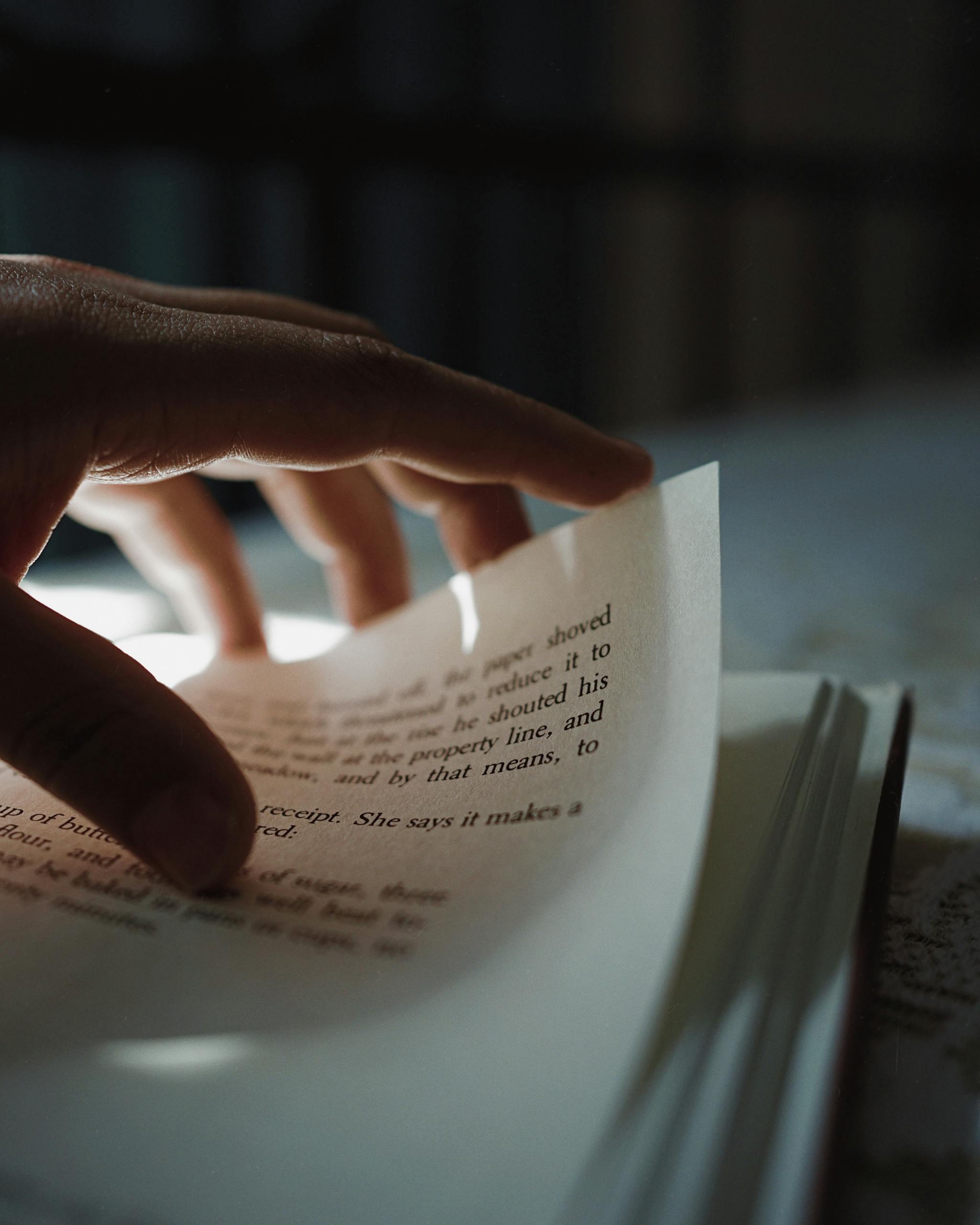 Close-up of a hand turning a page in an open book with blurry text in dim light.