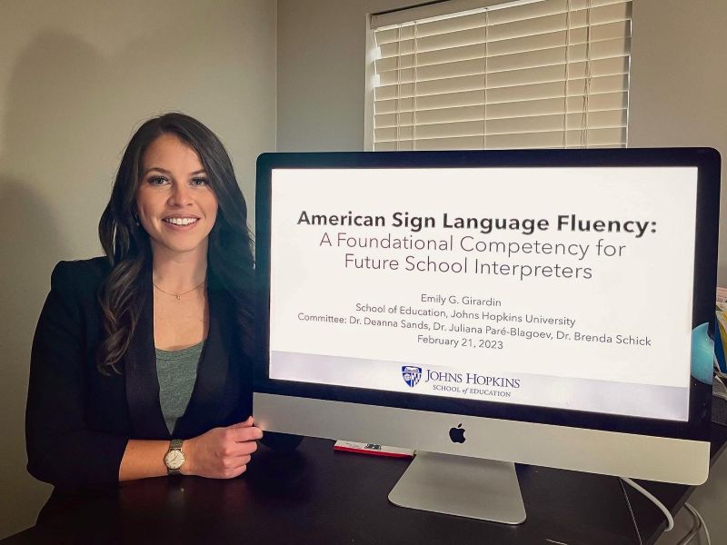 Dr. Emily Girardin stands beside a screen displaying her Johns Hopkins University dissertation presentation, “American Sign Language Fluency: A Foundational Competency for Future School Interpreters.”