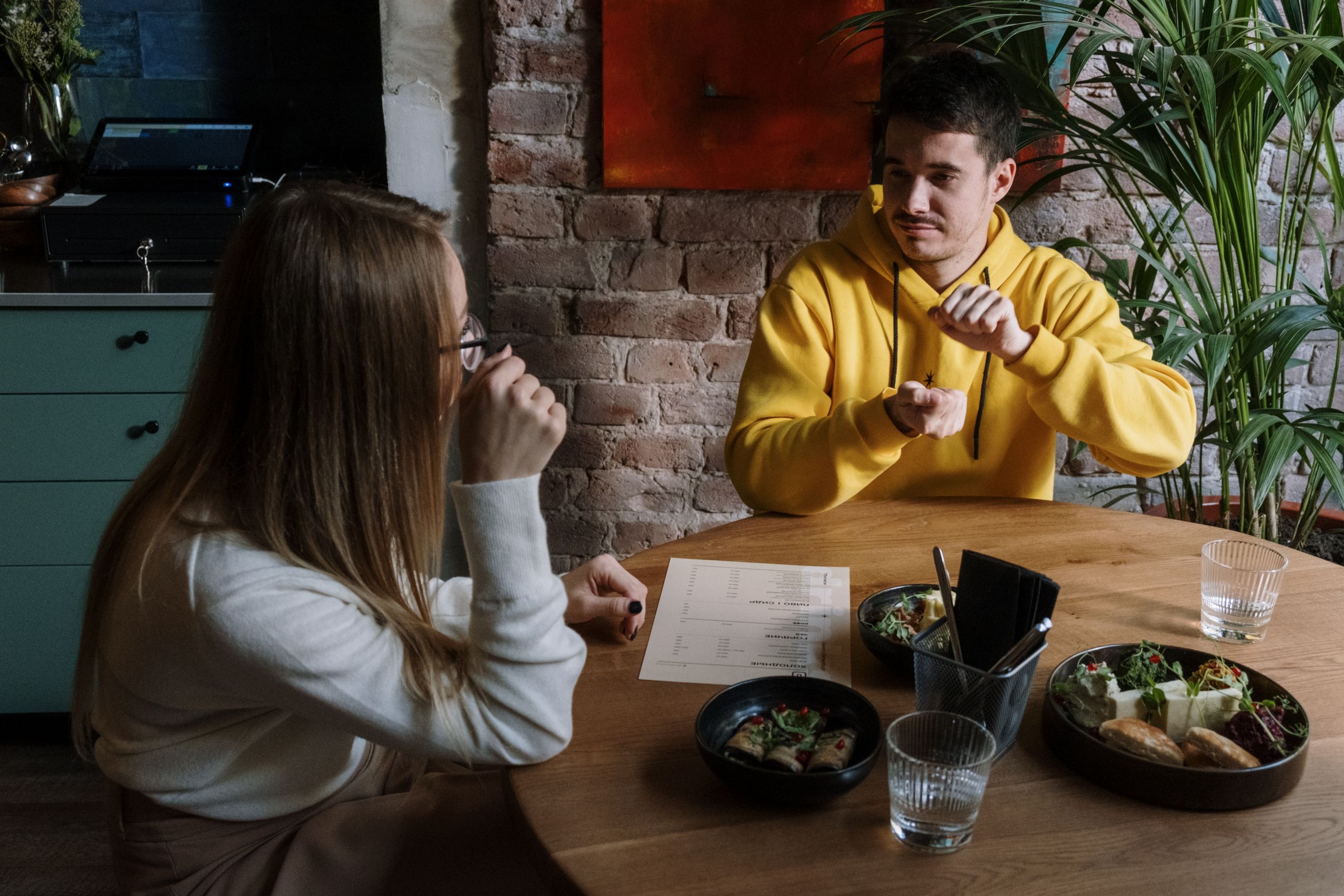 Two friends communicate in sign language over a meal in a cozy café setting.