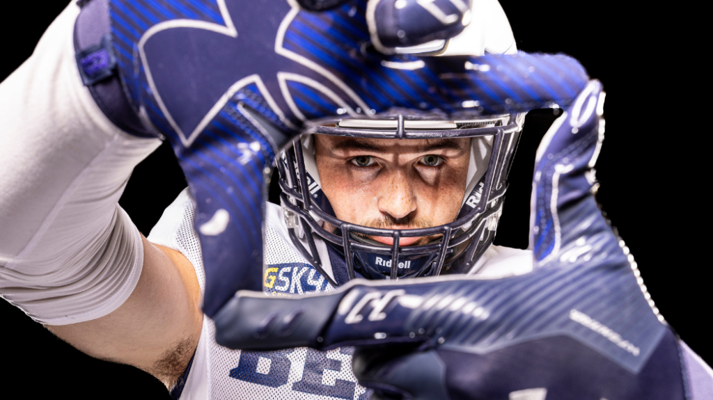A close-up of a UNC football player's face with his gloves making a square in front of his helmet