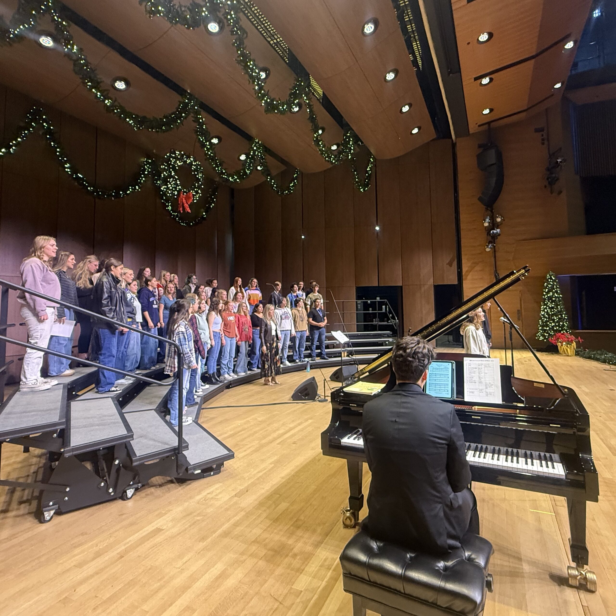 9th Ave Voices choir singing on stage in UNC Campus Commons Performance Hall with piano.