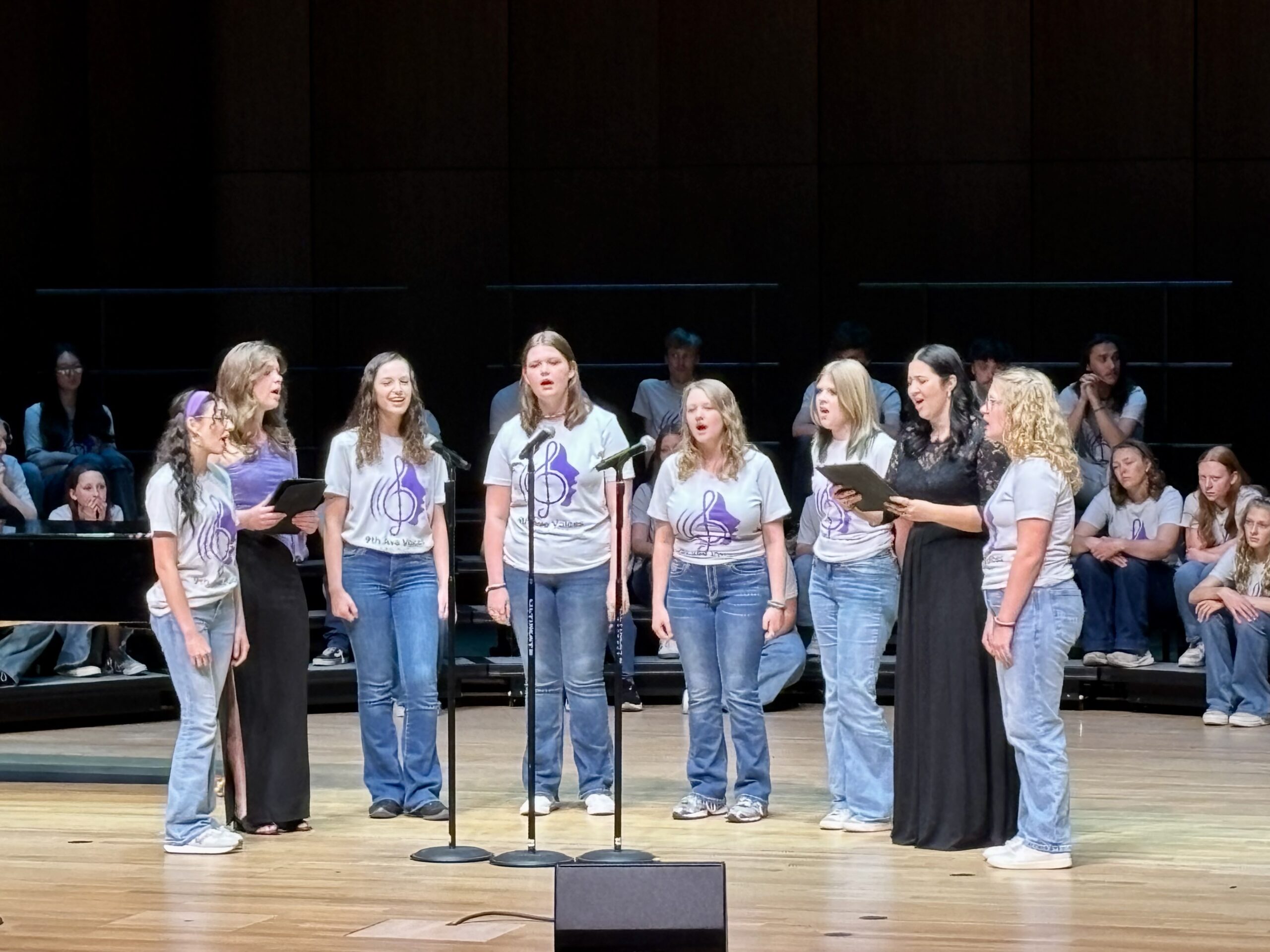A small group of singers singing into microphones on stage with 9th Ave Voices.