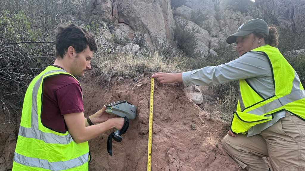 Two UNC students wearing yellow construction vests while holding an XRF device and a measuring tape against rock