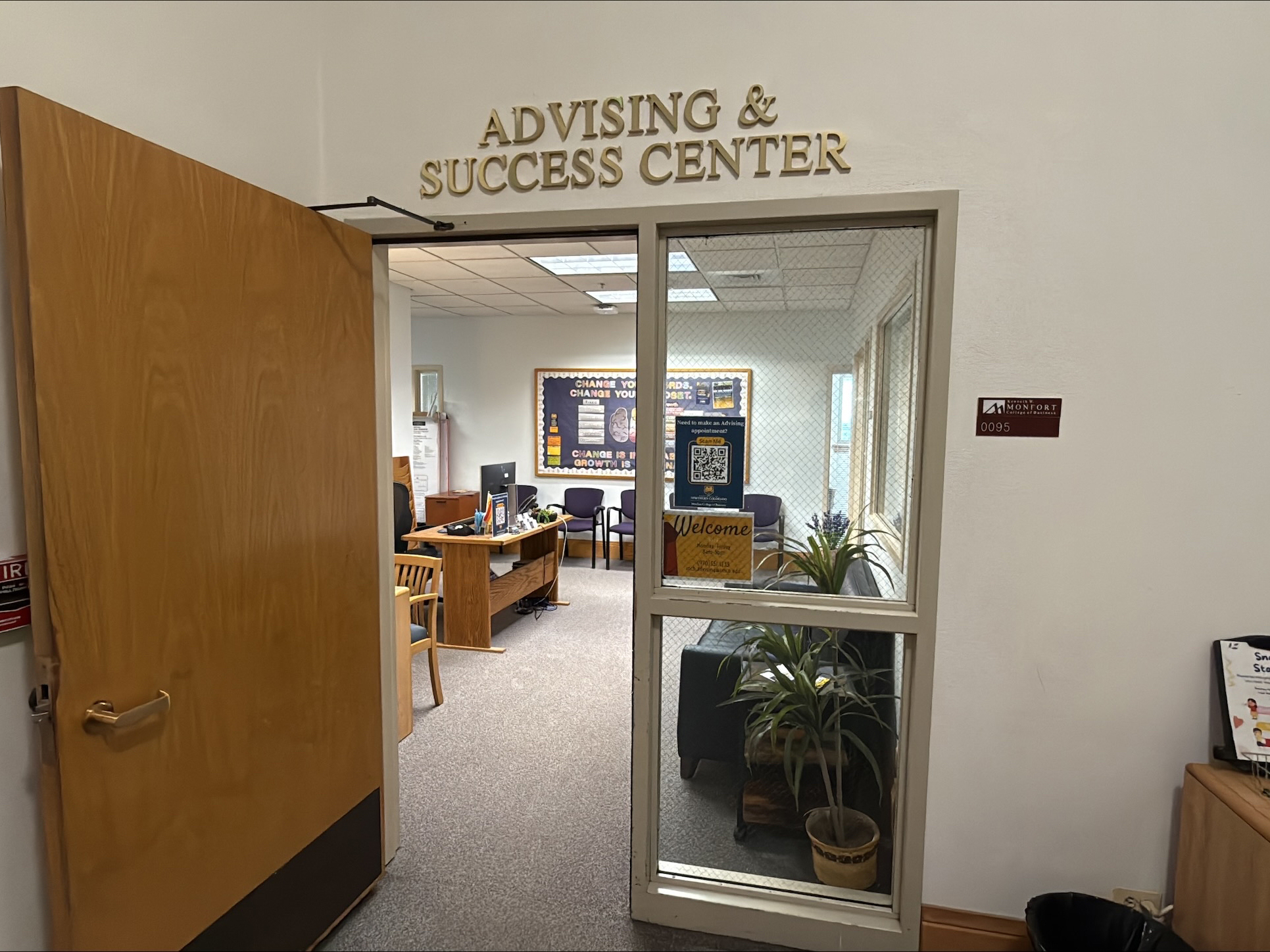 A glass door leading to the Advising & Success Center, showing a welcoming office with chairs, a bulletin board, and small plants. The office has a sign with 