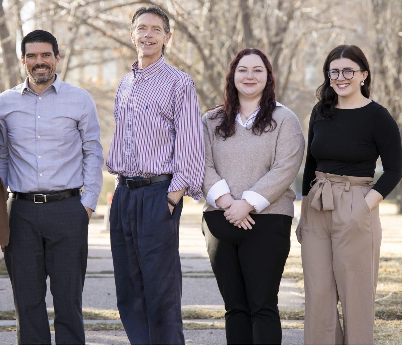 A group of four individuals from the Monfort College of Business advising team standing outdoors. They are dressed in professional attire and are smiling at the camera. The background features leafless trees, suggesting it is fall or winter.