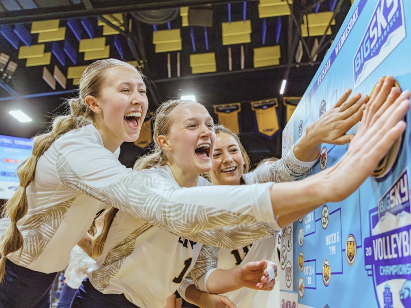 Three UNC Bears volleyball players touch a banner at the Big Sky championship in 2025