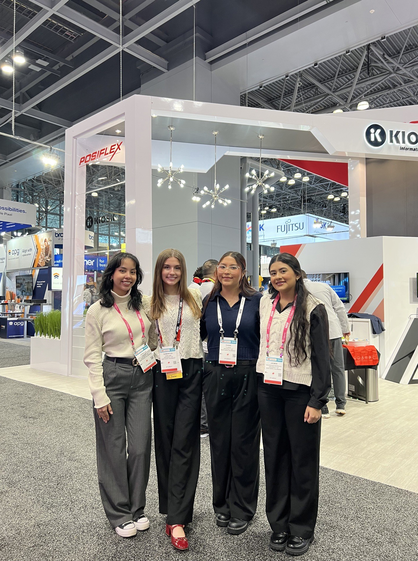 Four students pose together at a trade show in a large convention center, standing in front of a modern exhibition booth. They are dressed in professional attire, smiling, and appear to be engaged in the event. The booth features branded signage and an open, inviting design.