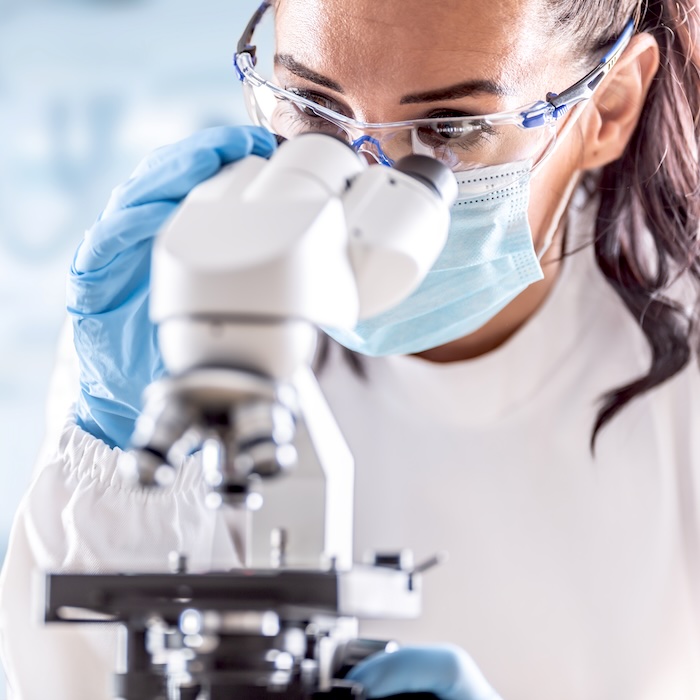 Female lab technician in protective glasses, gloves and face mask sits next to a microscope in laboratory.