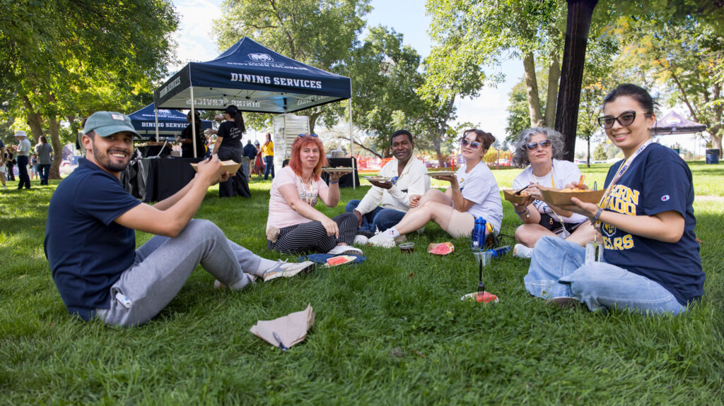 A group of six people sitting on the ground eating food outside
