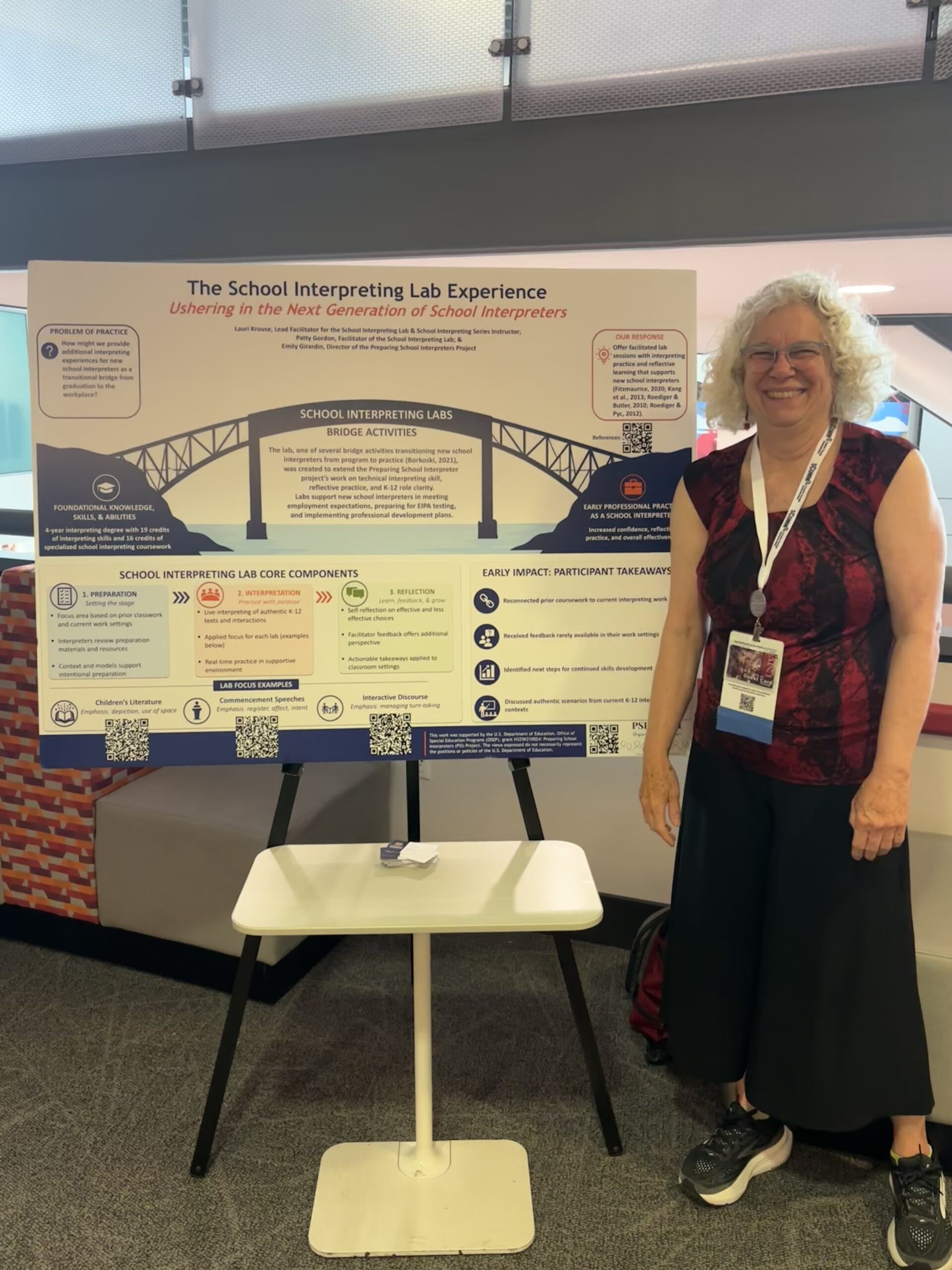 Lauri Krouse standing beside The School Interpreting Lab Experience poster at the 2026 NAIE Conference.