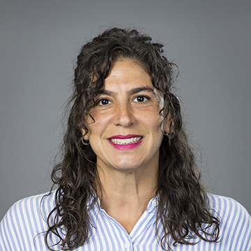 A person with long, dark, wavy hair smiles while wearing a blue and white striped shirt against a plain gray background.