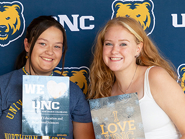 Two smiling students standing in front of a blue backdrop with UNC and bear logos, each holding a sign: the left sign reads 