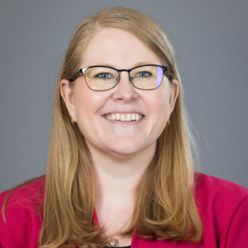 A smiling person with long blonde hair and glasses wears a bright pink blazer over a black top against a gray background.