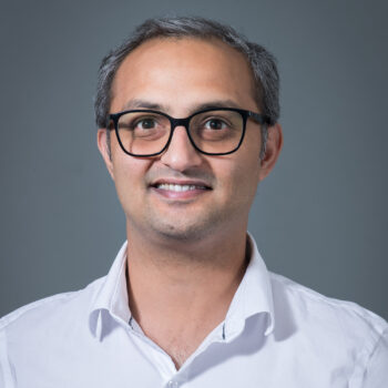Head-and-shoulders portrait of a smiling man with short salt-and-pepper hair and black glasses, wearing a white collared shirt against a plain gray background.