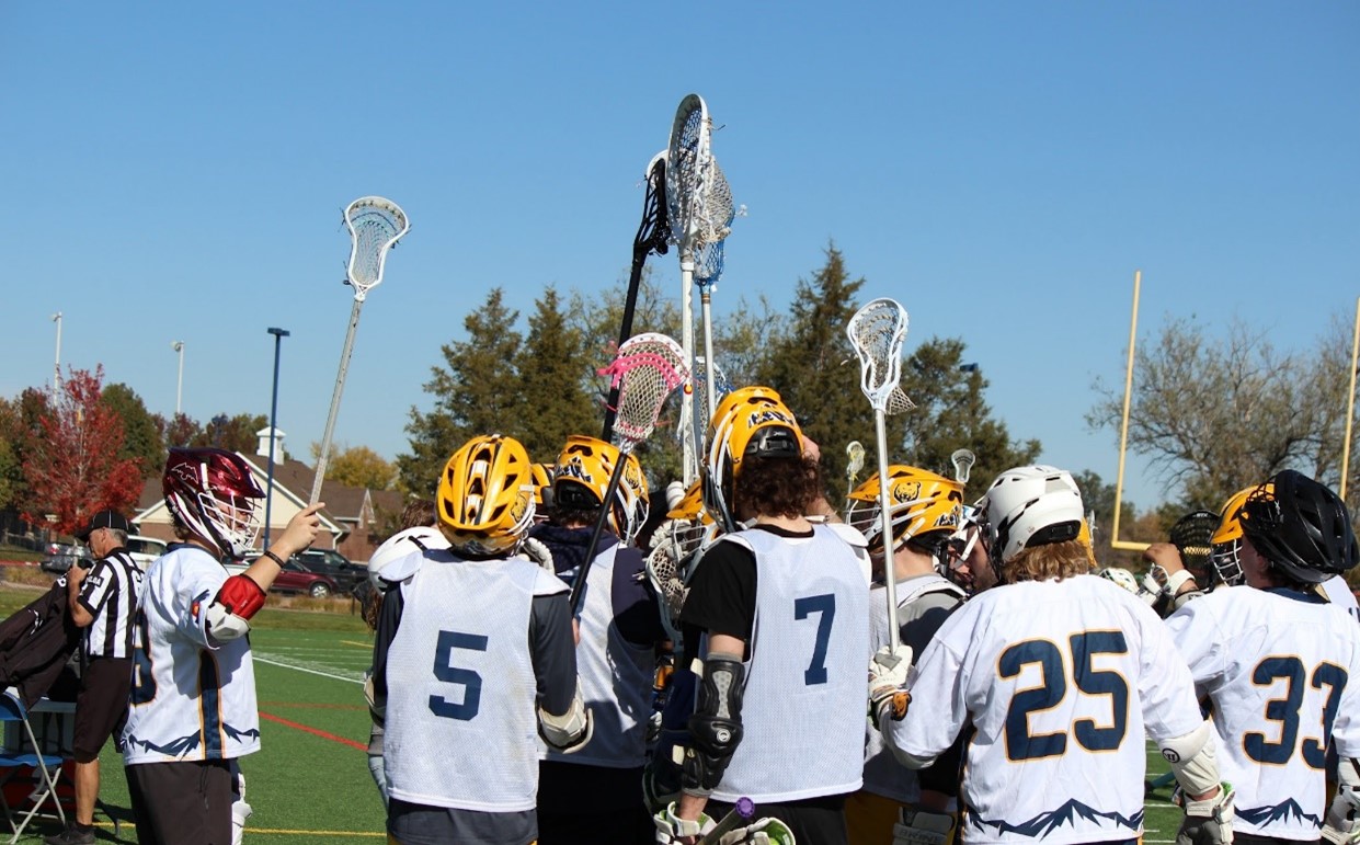 Photo of the Men's Lacrosse team in a huddle on the field raising their sticks in the air.